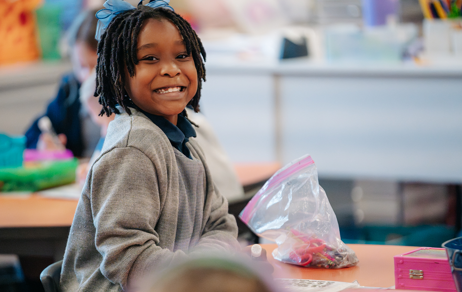 Smiling child at a classroom table with a bag of craft supplies