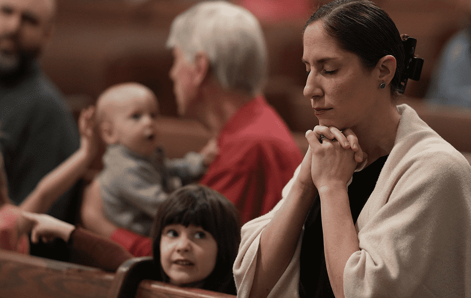 Woman praying with eyes closed in a church pew, with children and other congregants in the background