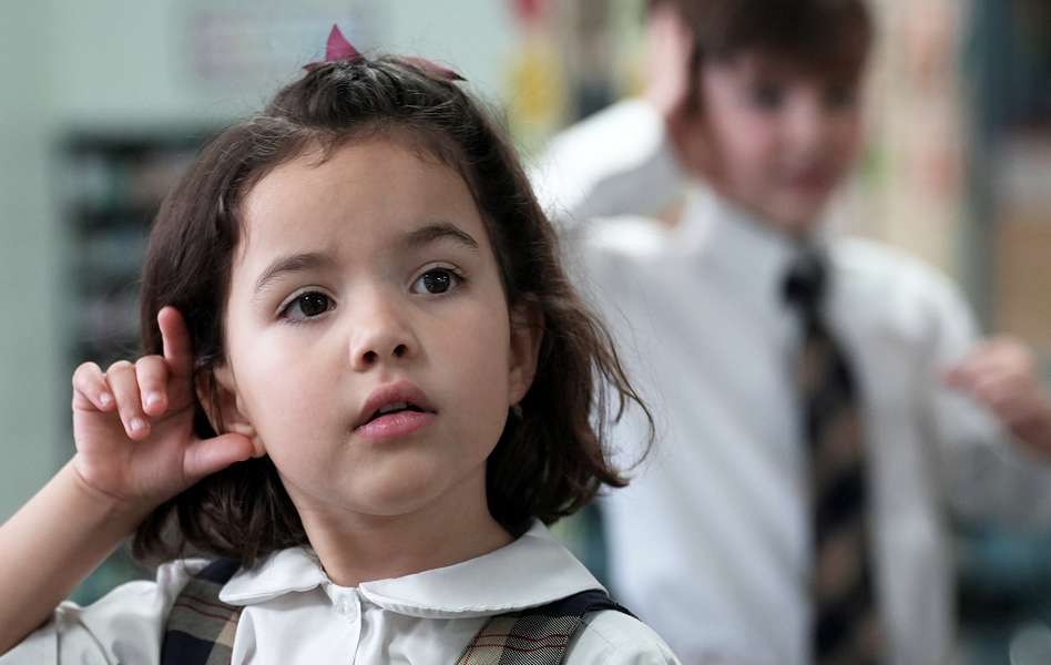 Young girl holding a hand to her ear in a classroom, with a blurred boy in the background