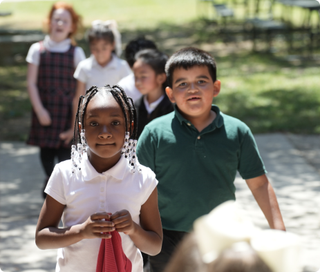 Children standing outdoors in a schoolyard, with two children in the foreground holding hands with a red cloth