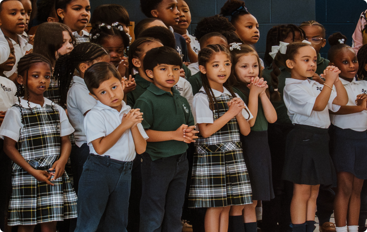 Children in school uniforms standing together with hands clasped, as if in prayer or singing at a school event