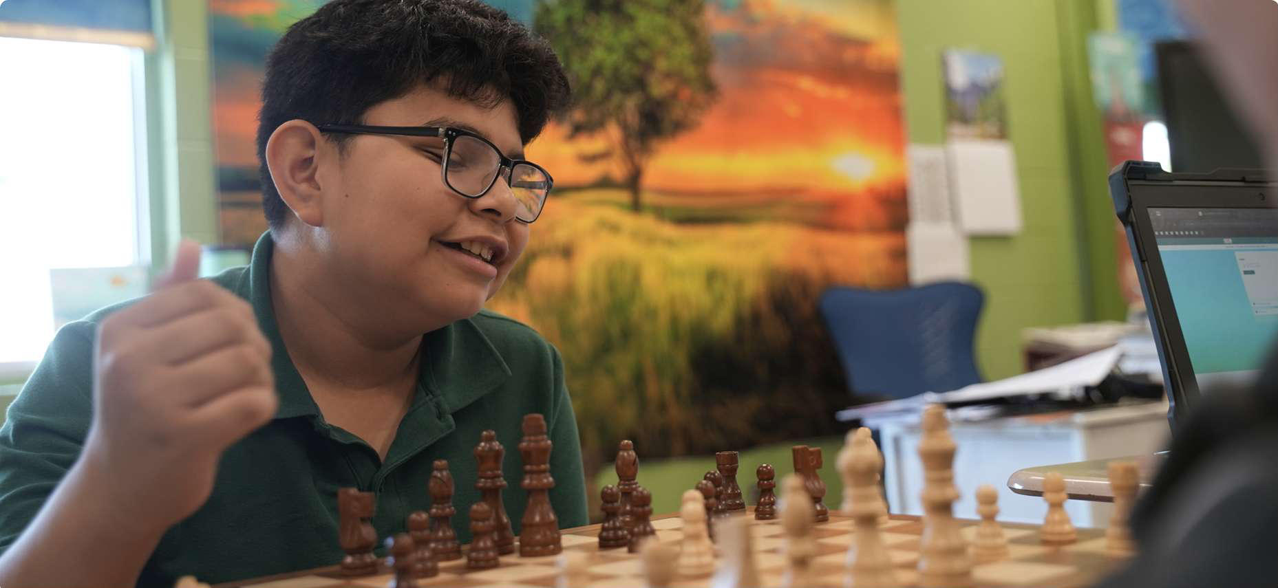 Boy playing chess at a table, studying the board with a laptop nearby