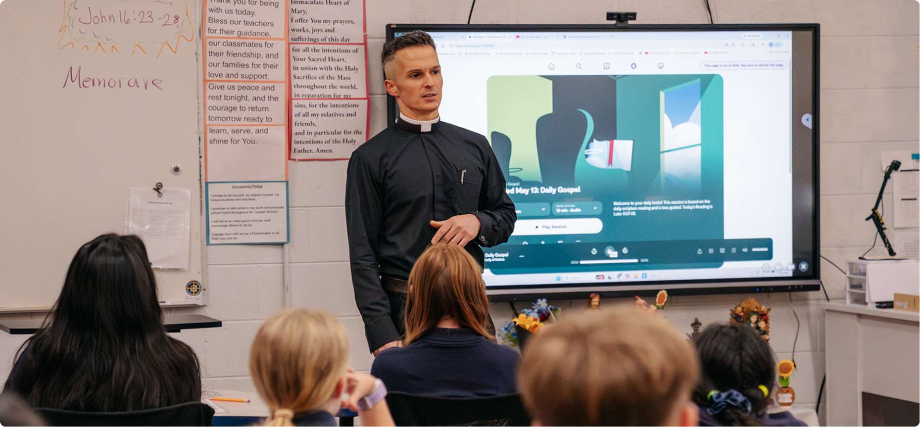 Catholic priest speaking to students in a classroom with a scripture lesson displayed on a screen