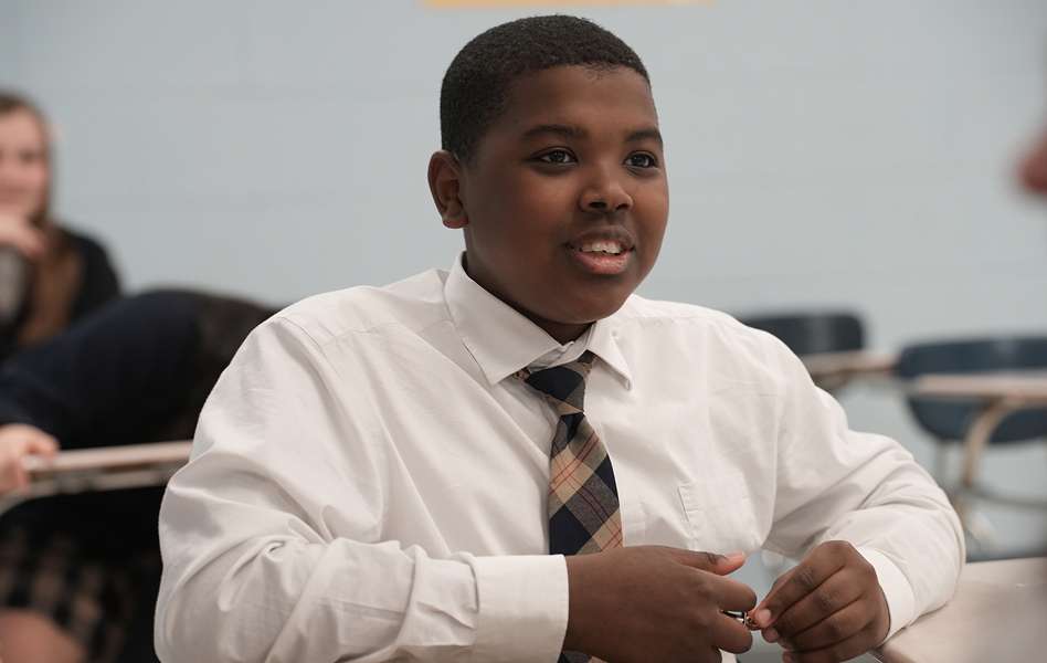 A student in a white shirt and tie sits at a desk in a classroom