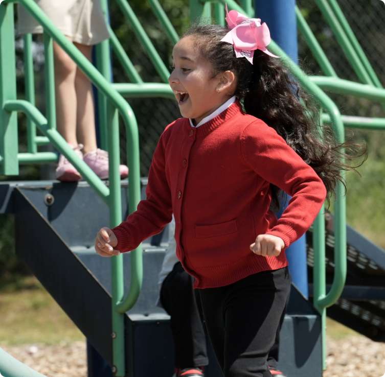 A young girl in a red sweater runs down playground steps with a pink bow in her hair