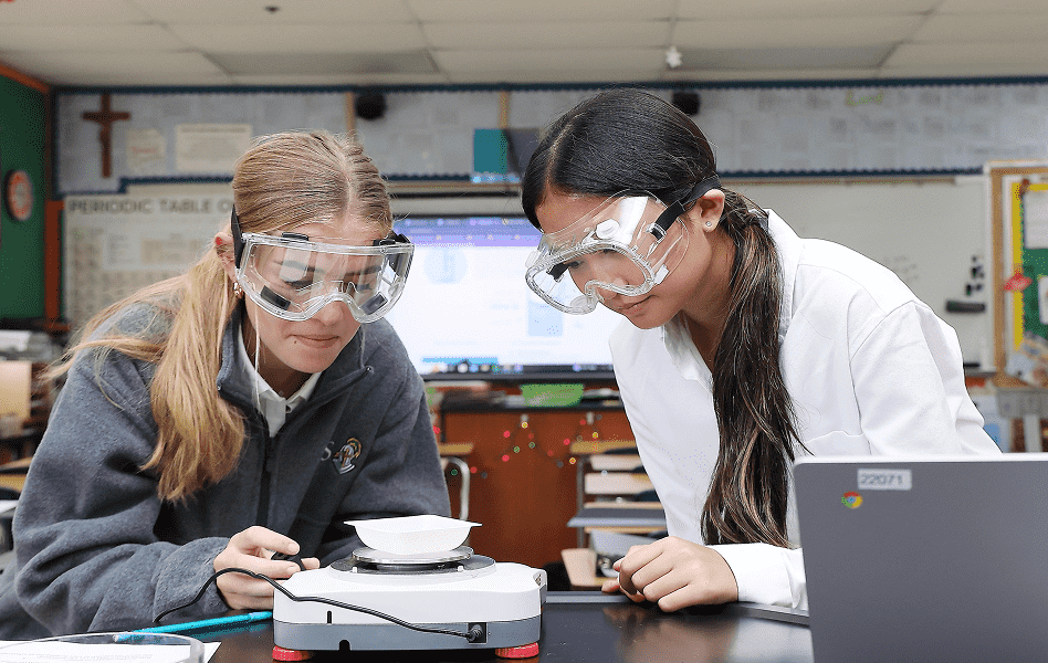 Two students wearing safety goggles examine a small dish on a lab hot plate in a classroom