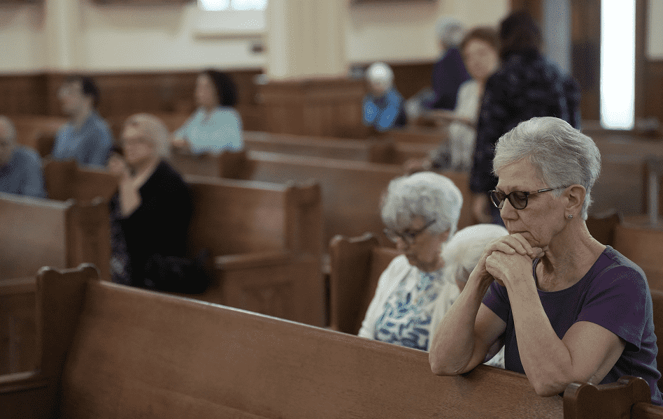 An older woman sits in a church pew with her hands clasped in prayer
