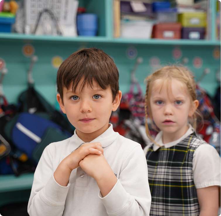 Two children standing in a classroom, with backpacks hanging on the wall behind them