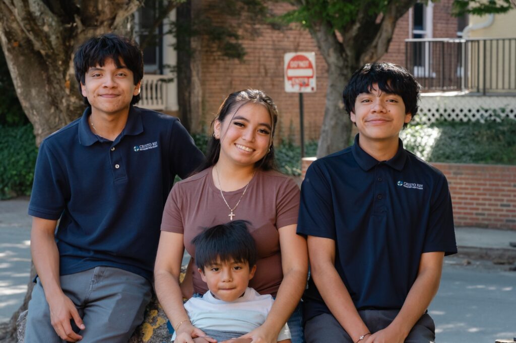Three smiling teens and a young child sitting outdoors in front of a brick building